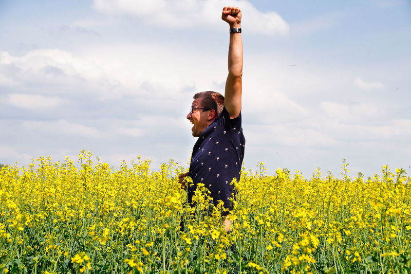 Happy mid adult man with arms raised standing on oilseed rape field against cloudy sky