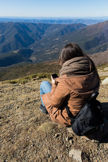 Rear view of woman using phone while sitting on mountain during winter