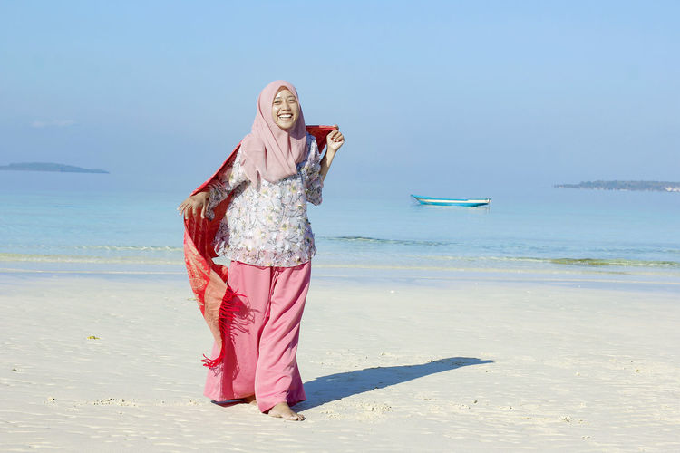 Smiling young woman wearing hijab walking at beach against clear blue sky during sunny day