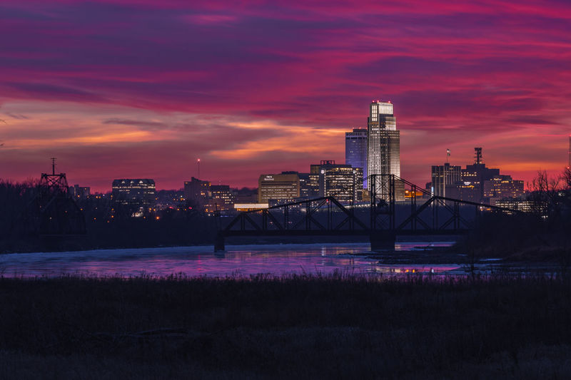 Illuminated buildings by river against sky during sunset