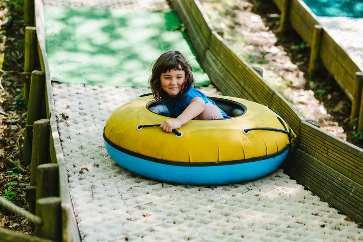 Side view of delighted little child in t shirt smiling while riding inflatable sled tube on slide on playground in sunny adventure park