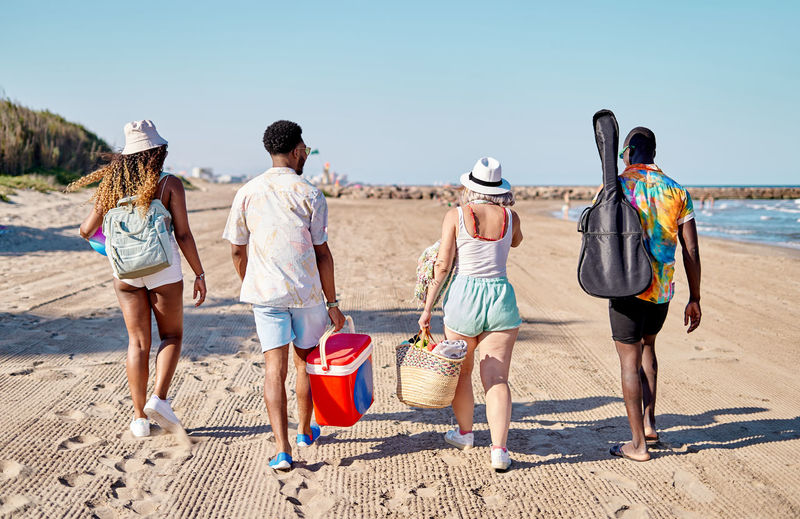 Full body back view of unrecognizable multiracial men and women in sunglasses and hat strolling on sand against cloudless blue sky while spending time on beach on summer day