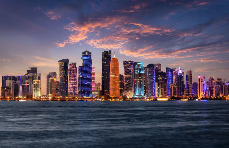 Sea by illuminated buildings against sky at sunset