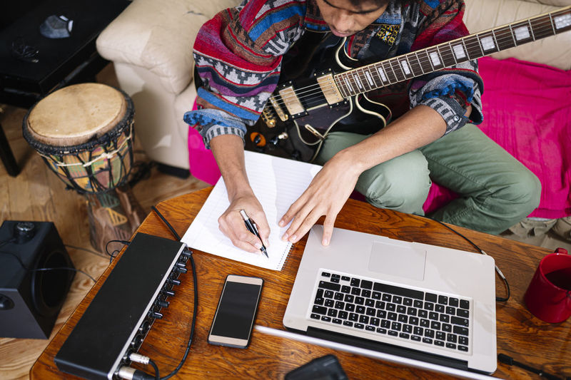 Young man with guitar writing notes on book by laptop in living room
