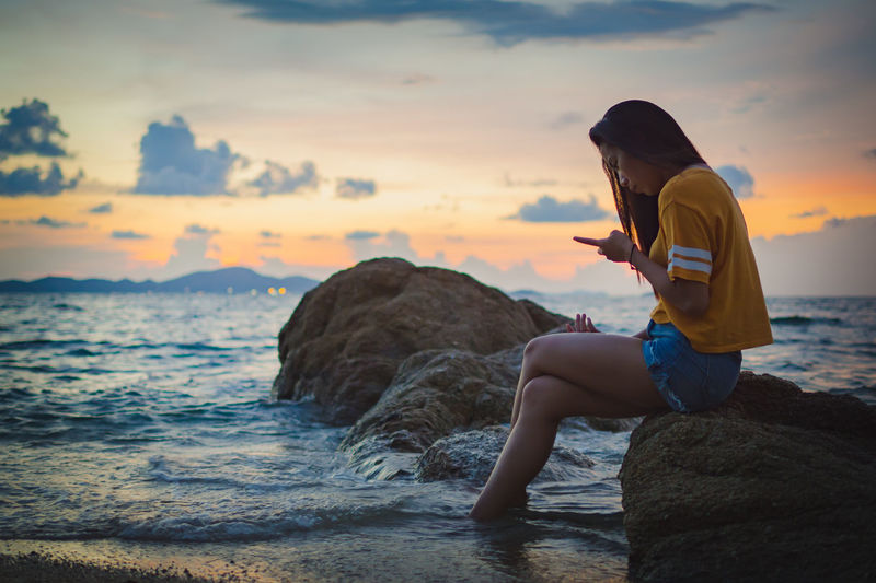 Side view of woman sitting on rock at sea against sky during sunset