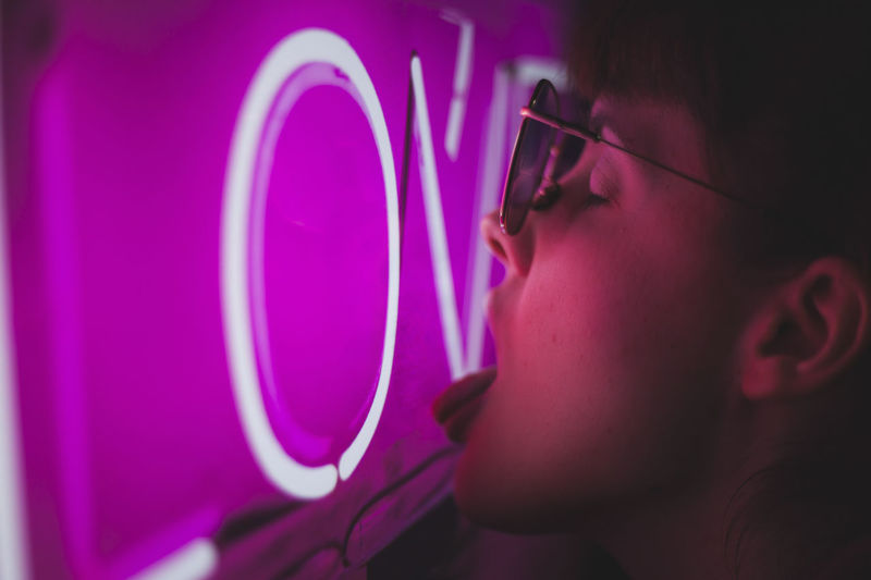 Side view of expressive young stylish female licking pink neon sign glowing in dark in night city