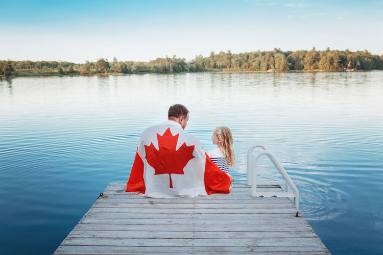 Father and daughter wrapped in canadian flag sitting on pier by lake. canada day celebration outdoor