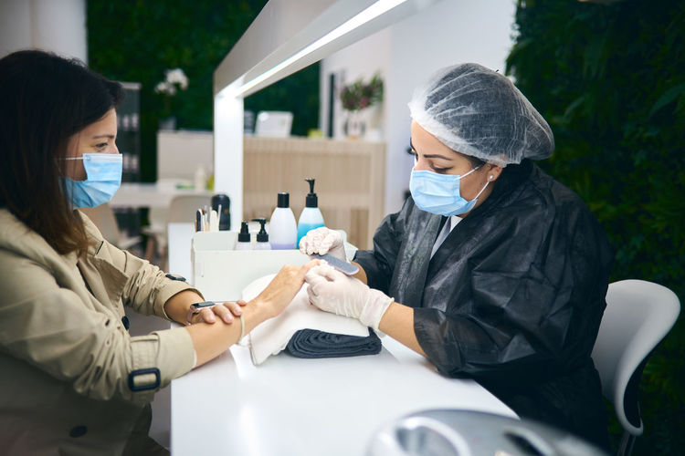 Side view of cheerful manicure master using nail file while sitting at table with female client in modern beauty salon during coronavirus pandemic