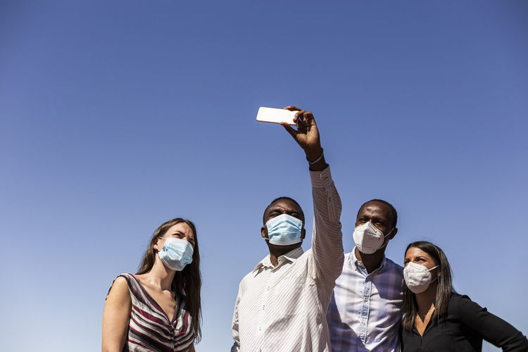 Business professionals taking selfie with protective face masks against clear blue sky on sunny day