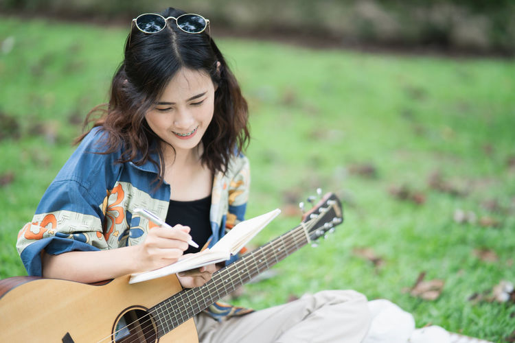 Young woman writing in diary while holding guitar while sitting at lawn