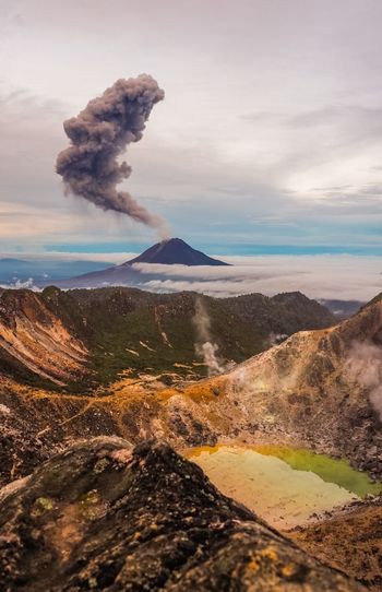 Smoke emitting from mountain against cloudy sky