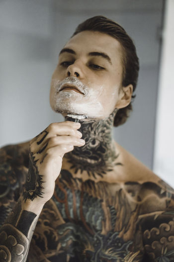 Shirtless tattooed man shaving in bathroom