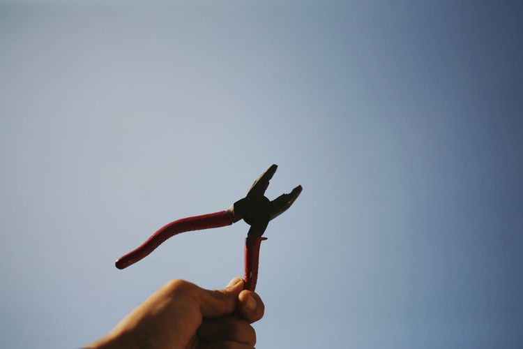 Low angle view of person hand holding pliers against clear sky