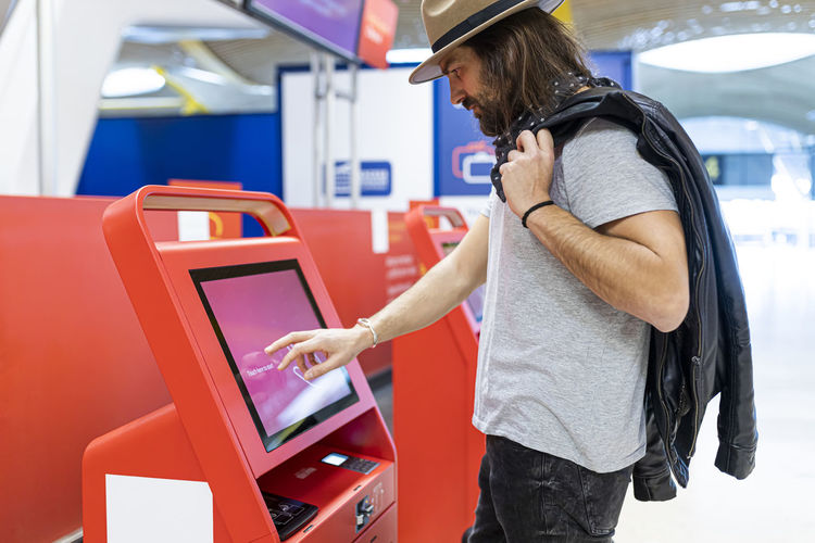 Side view of bearded man with long hair and hat caring a suitcase using self check in in a modern machine in a airport terminal
