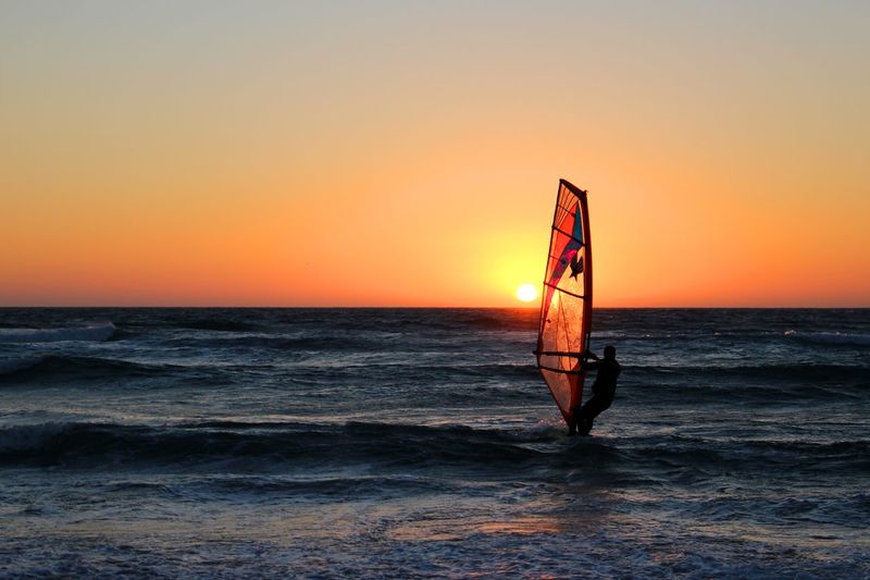 Silhouette man windsurfing on sea against orange sky