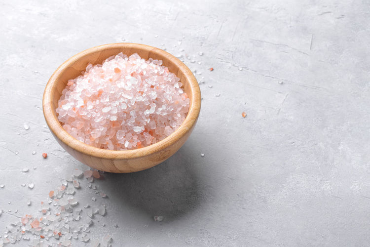 Pink himalayan salt in a wooden bowl with scattered salt on a light grey background, close up