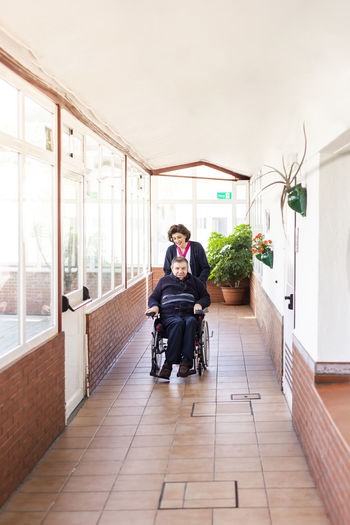 Female nurse helping disabled man sitting on wheelchair in corridor of nursing home