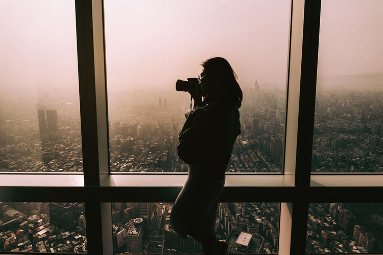 Woman photographing through window against cityscape