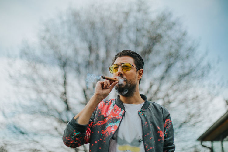 Young man smoking cigar while standing against sky