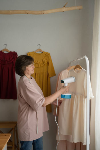 Mature woman steaming handmade white dress hanging on rack during work in light workshop in daytime