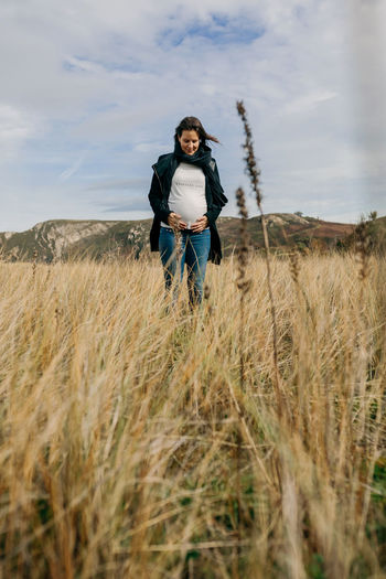 Pregnant young woman touching abdomen while standing on grass against sky