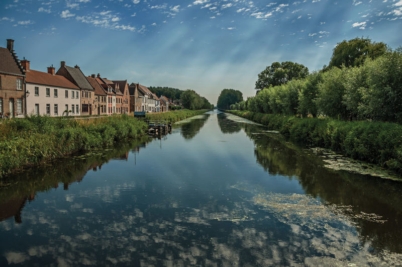 Trees and houses along canal with sky reflected on water in damme. a country village in belgium.