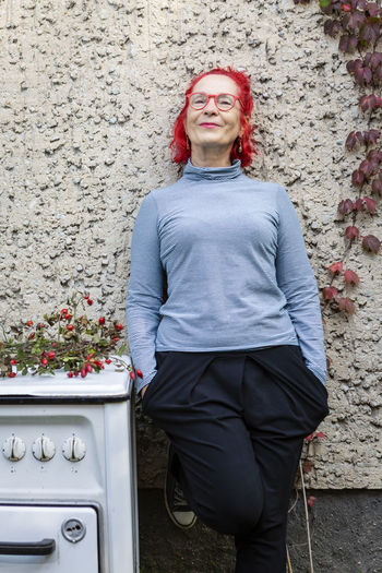 Portrait of smiling senior woman with red dyed hair leaning against house front