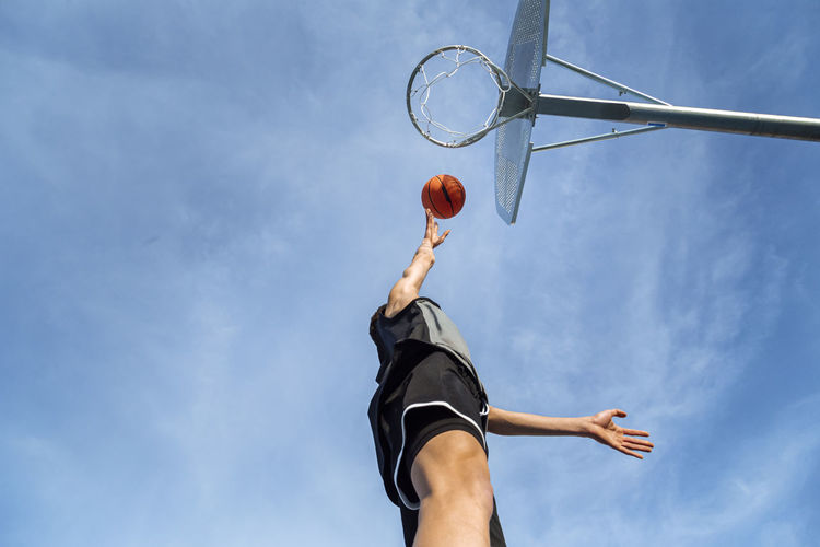From below of anonymous sportive male throwing ball into basketball hoop while playing game against blue sky on sunny summer day