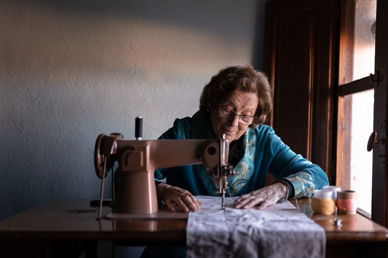 Senior female sewing white textile on special machine at table with spools of thread in room