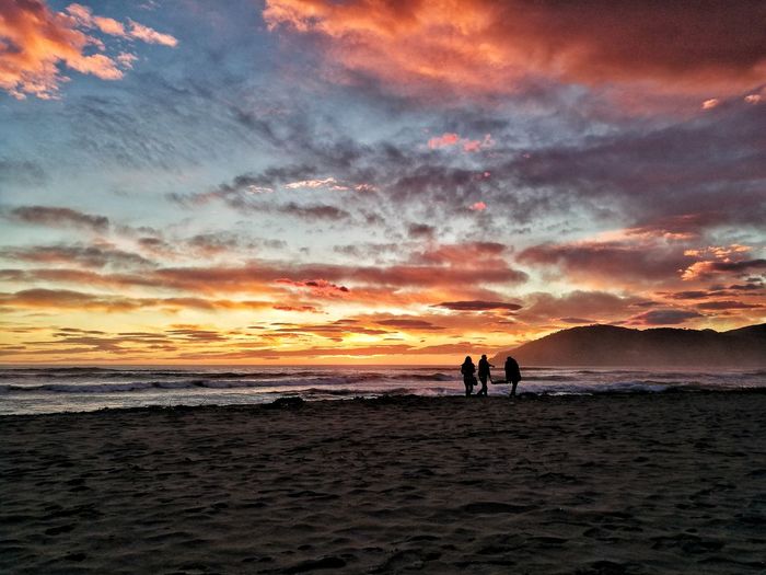 Silhouette people on beach against dramatic sky during sunset