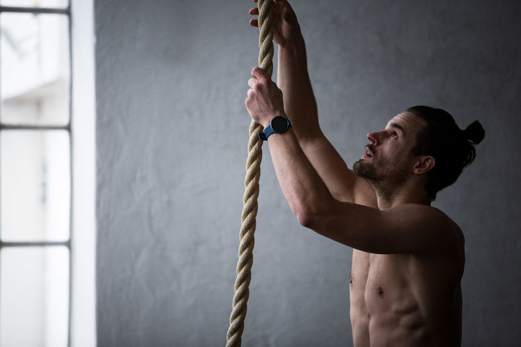 Side view of shirtless man holding rope against wall