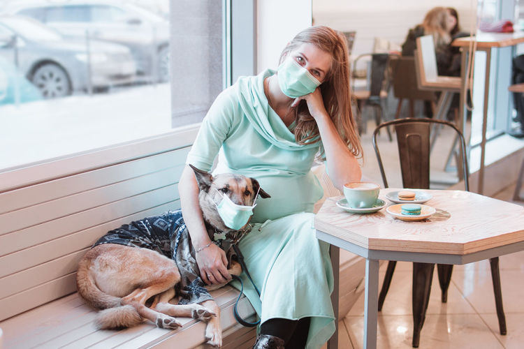 Pregnant women and her dog in protective mask are sitting at the table in cafe. 