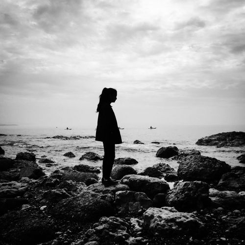 Side view of silhouette teenage girl standing on rocks at beach against sky