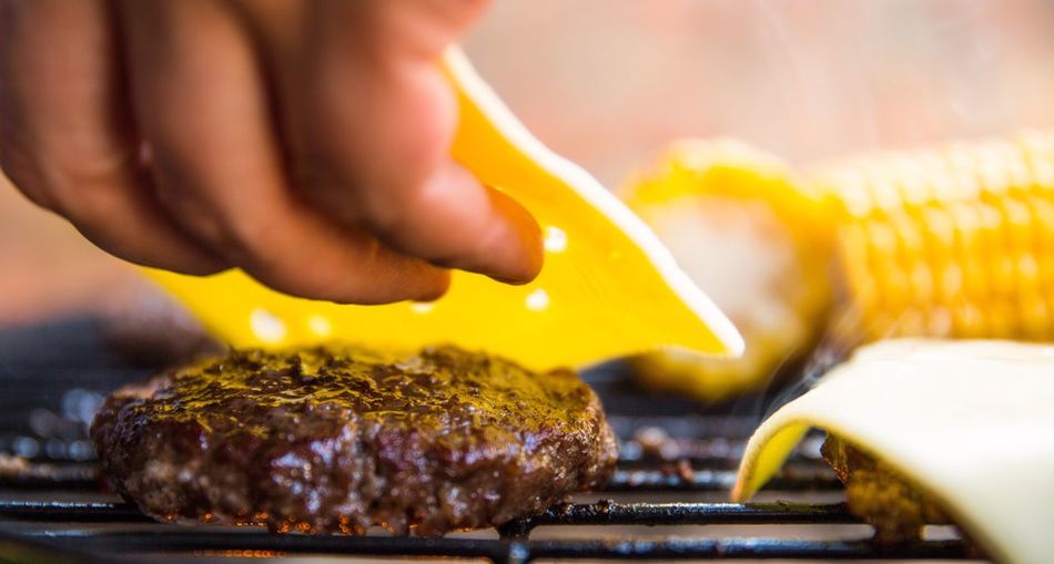 Cropped image of person placing cheese over burger on barbeque grill