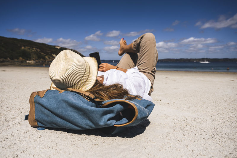 Mature woman wearing hat using smart phone lying at beach