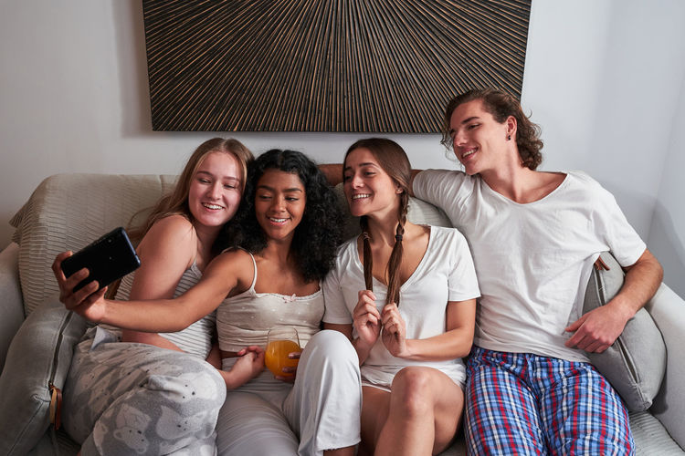 Full body excited young multiracial flatmates in domestic clothes taking selfie on smartphone while gathering in modern living room