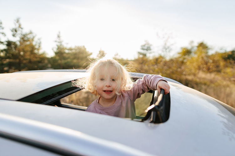 Adorable girl with blond hair looking at camera with smile while standing in open car sunroof during road trip in countryside