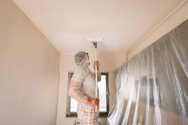 Man in protective workwear and mask working with paint roller on wall in kitchen at home