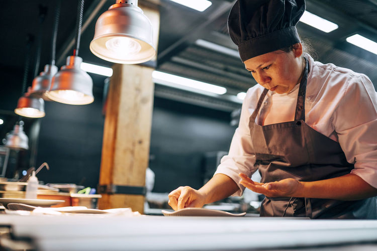 Adult woman in apron and hat preparing and serving dishes on kitchen in modern restaurant
