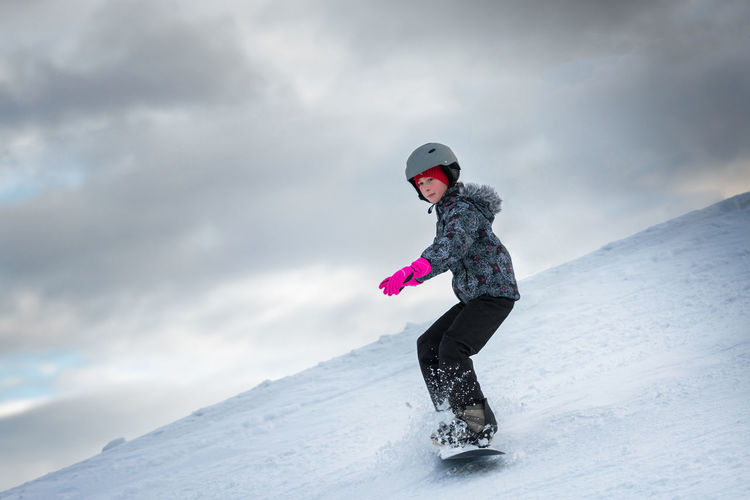 Full length of girl snowboarding on snowy mountain against cloudy sky