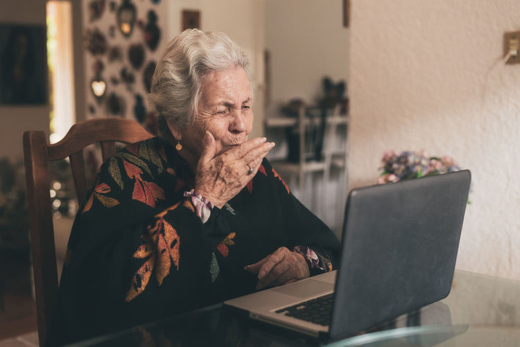 Positive elderly female wearing warm clothes sitting at table and sending air kiss while talking by video call using laptop
