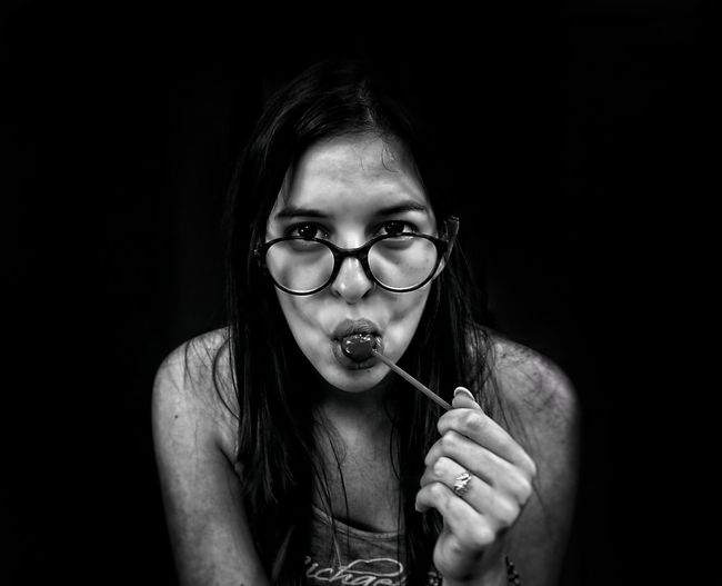Portrait of beautiful young woman eating lollipop against black background