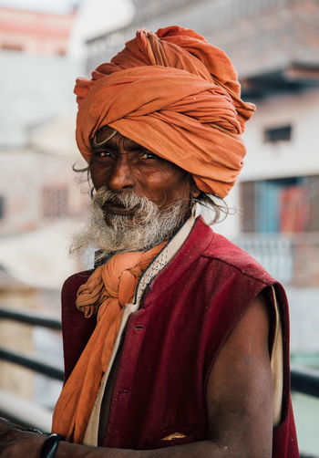 Varanasi, india - february, 2018: aged bearded hindu male in orange turban wearing red sleeveless vest standing on street and looking at camera