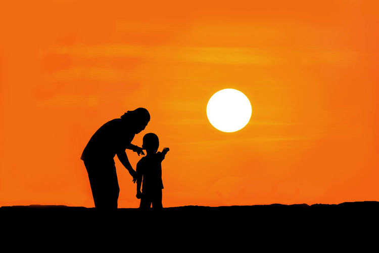 Black silhouette of a mother and son watching the sunset on mountain orange background
