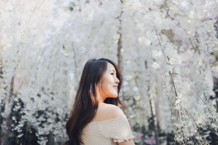 Thoughtful young woman smiling while standing against white flowering trees at park