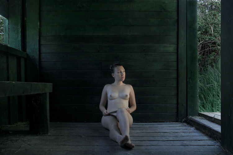 Full length of naked woman sitting by wooden wall