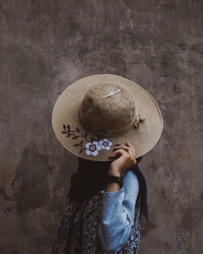 Young woman hiding face with hat against wall