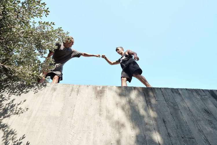 From below of friendly men greeting each other and bumping fists while preparing for parkour training on stone building in city