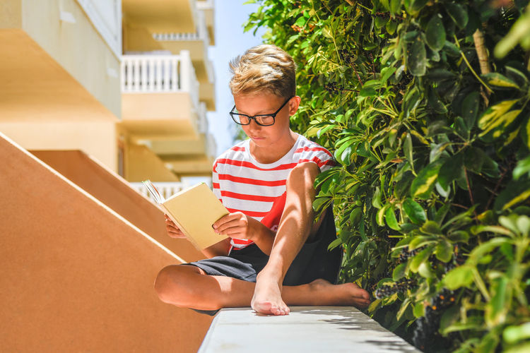 Full length of boy reading book while sitting on building terrace