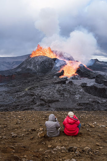 Back view of unrecognizable travelers admiring fagradalsfjall with fire and lava while taking photo and sitting on mount in iceland